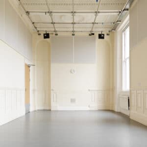 A long picture of a white room. Hanging from the ceiling is a lighting rig and there are two big windows against the right side wall.