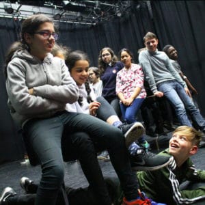 A group of teenagers acting in a black room. two groups are leaning on a table. A girl sits on a chair with her arms folded and looks angry while a boy lies on the floor smiling.