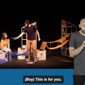 A BSL performer on the right signing along to the video of Paper Aeroplane. There are two actors, a girl in a yellow t-shirt sits on a box and a man in a black t-shirt stands. They are both wearing flight goggles. The male actor hands something to the female and says 'This is for you.'