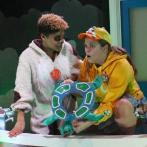 Two actresses in a big red bath. One is dressed as a dog and the other as a duck. They hold a blow up turtle and there are two toys of a lion and giraffe in the bath too.