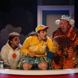 Three actors dressed as a dog, duck and lion are in a big red bath looking at something in the palm of the hand of the duck character