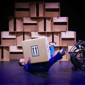 Daryl, a white man wearing a blue overall lays on his back the floor with a large brown cardboard box on top of his chest. His arms and legs are stretched out. Next to him is his upturned wheelchair. In the background is a wall of boxes.