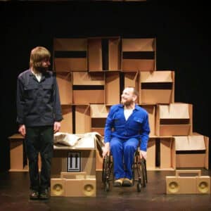 Daryl, a white man wearing a blue overall sits in his wheelchair. Next to him stands Jon, A white man wearing a dark blue overall. They are looking at each other. On the floor around them are radios mad out of cardboard and behind them is a wall of boxes.