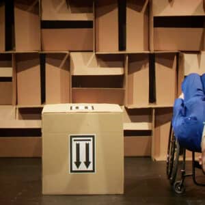 Daryl, a white man wearing a blue overall sits upside down in his wheelchair, his head almost resting on the floor as he looks outwards. Next to him is a large cardboard box with 2 arrows on it pointing to the floor. In the background is a wall of boxes.
