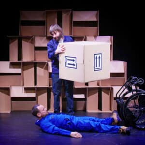 Daryl, a white man wearing a blue overall lays on his back the floor next to his upturned wheelchair. Jon, A white man wearing a dark blue overall, stand over him holding a large cardboard box. In the background is a wall of boxes.