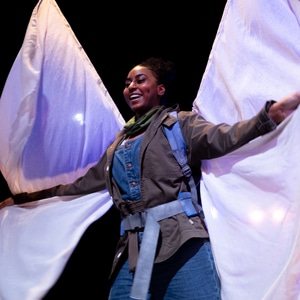 A woman standing wearing white butterfly wings made out of white fabric. She is smiling and holding the wings out.