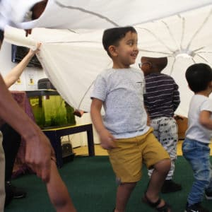 Three little boys run underneath a white parachute that their parents are holding up.