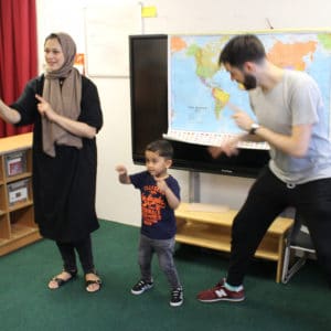 A woman and man stand in front of a world map in a classroom. A little boy stands in between them and they are all pointing.