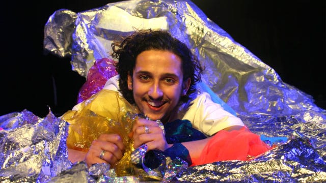 A male actor with dark hair lies on the floor surrounded by shiney silver fabric and colourful plastic. He is smiling.