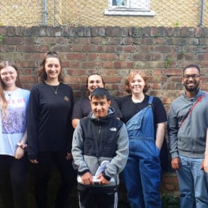 A photo of 8 young people standing in a line against a brick wall in a garden.
