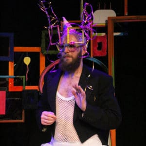 A man wearing a tutu and blazer is waving. He has multicoloured glasses on and a party hat.