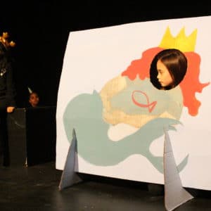 A little girl pokes her head through a cardboard cut out of a mermaid with red hair, a yellow crown and a green tale. Another girl is sat on a black box wearing black clothes and a gold inflatable crown.