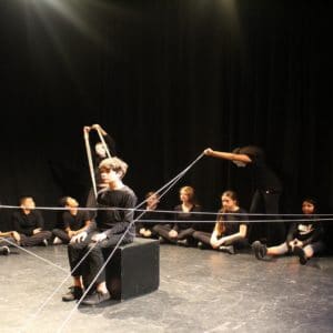 A dark room with a circle of young people dressed in black around the edge. Four young people with white masks stand and hold strings that are attached to one boy sitting in the middle on a black block.
