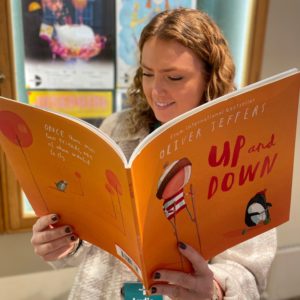 A woman with blonde curly hair is reading an orange book called Up and Down by Oliver Jeffers. The front cover of the book has a boy walking on stilts an a penguin. The woman is smiling as she reads the books.