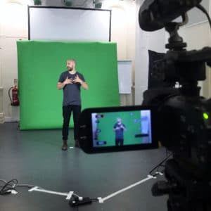 Through the lense of a film camera is a bald man standing in front of a green screen and doing sign language.