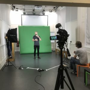 A white room with a green screen in the centre and a video camera in the foreground. A bald man stands in front of the greenscreen doing sign language and another man is sat on a chair watching from next to the camera.