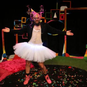 A photo of a man wearing a brightly coloured party hat, large glasses with streamers, a string vest, black jacket, tutu, and pink boots. He is standing with his arms outstretched looking straight out.