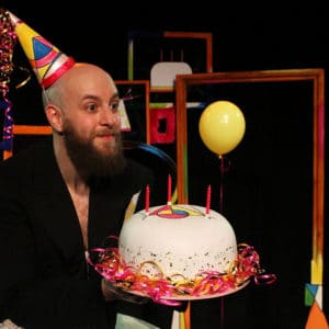A bald man with a beard holds a white birthday cake with three candles on it. He is earing a brightly coloured pointy party hat with streamers attached to the top. In the background are two yellow balloons.