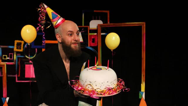 A photo of a man wearing a brightly coloured pointy party hat with streamers. He is holding a birthday cake with three candles in it.