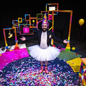 A male actor wearing a tutu, pink boots and a party hat standing on stage surrounded by confetti and balloons.