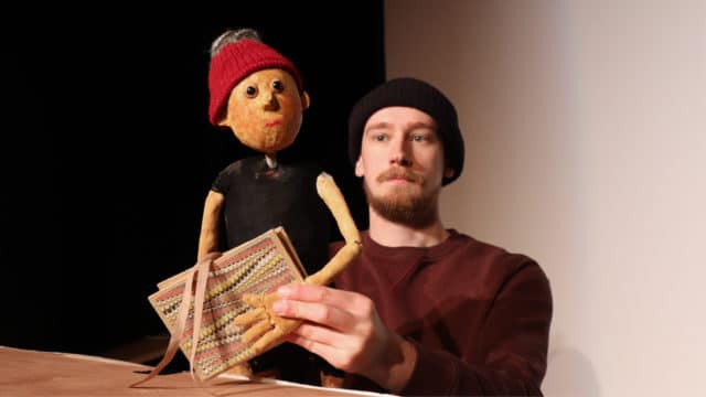 A man wearing a black beanie and red jumper with a beard and moustache holds a puppet of Pinocchio. The puppet has a red knitted hat and is holding a book.
